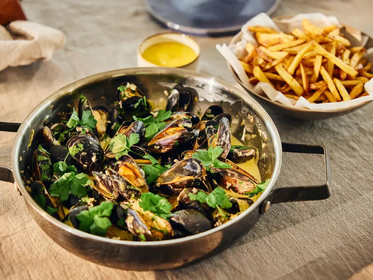 En stor kastrull med ångande moules frites, garnerad med persilja, bredvid en skål med gyllene pommes frites.