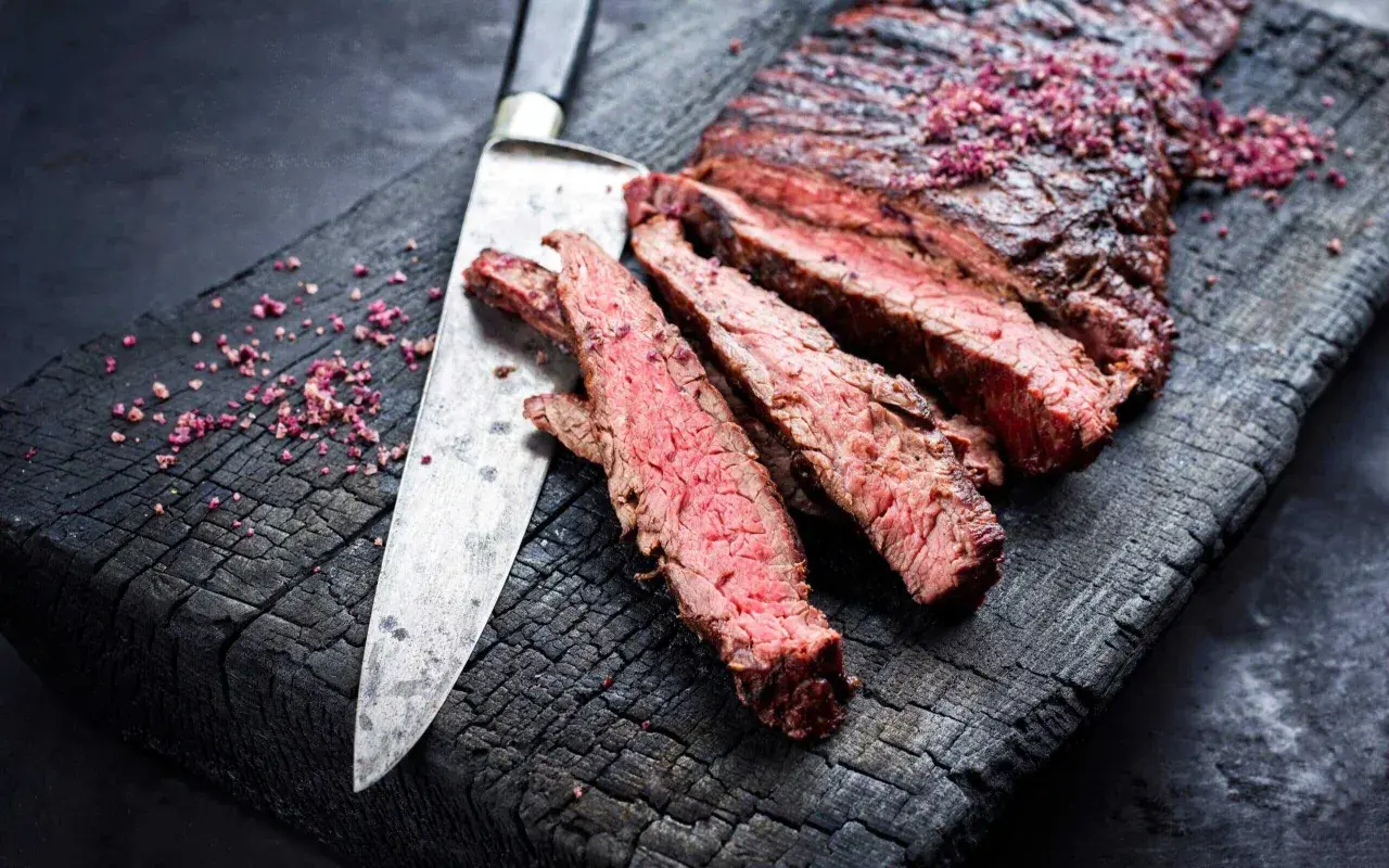 Skivat flap steak med rosa salt på en sotad träskärbräda, bredvid en kniv.
