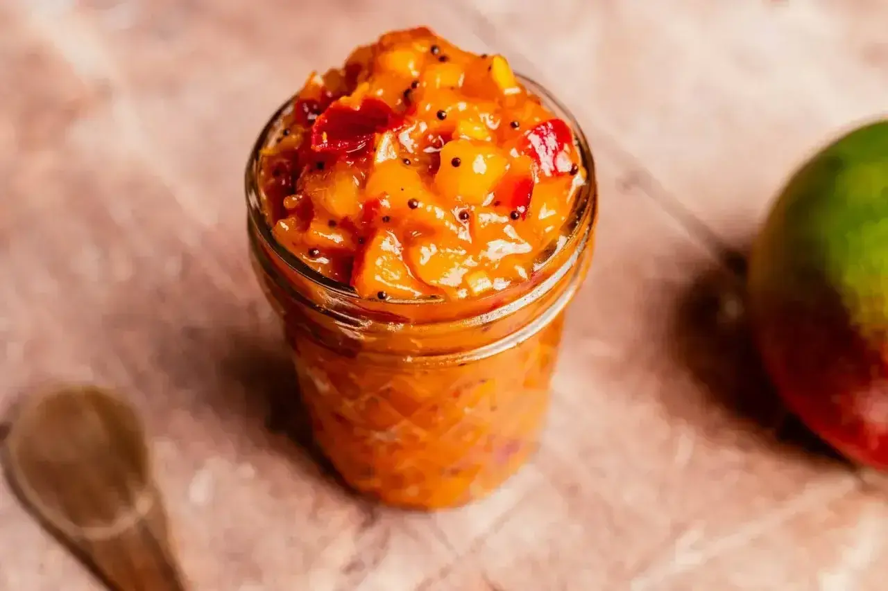 En burk med mango chutney, perfekt för ditt nästa chutney recept. En hel mango och en träsked syns också.