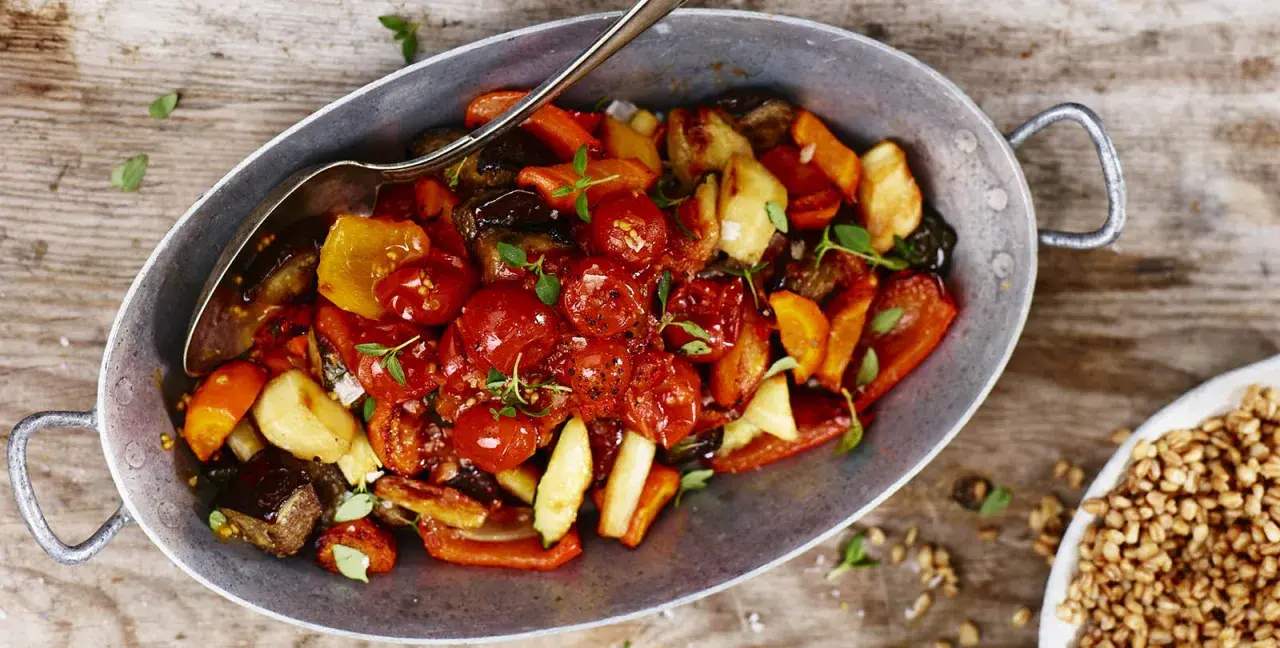 En varm ugnsbakad grönsaksblandning med körsbärstomater, paprika och aubergine, serverad i en metallskål med en sked. En härlig rostad tomatsås gör rätten extra smakrik.