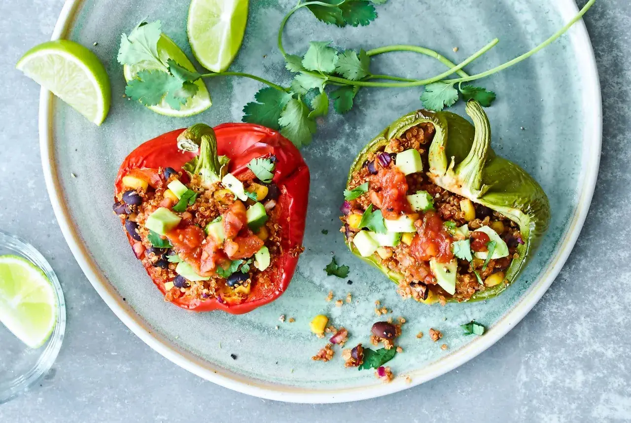 Två färgglada, fyllda paprikor med quinoa, svarta bönor och majs, toppade med salsa och avokado. Serveras med limeklyftor och koriander.