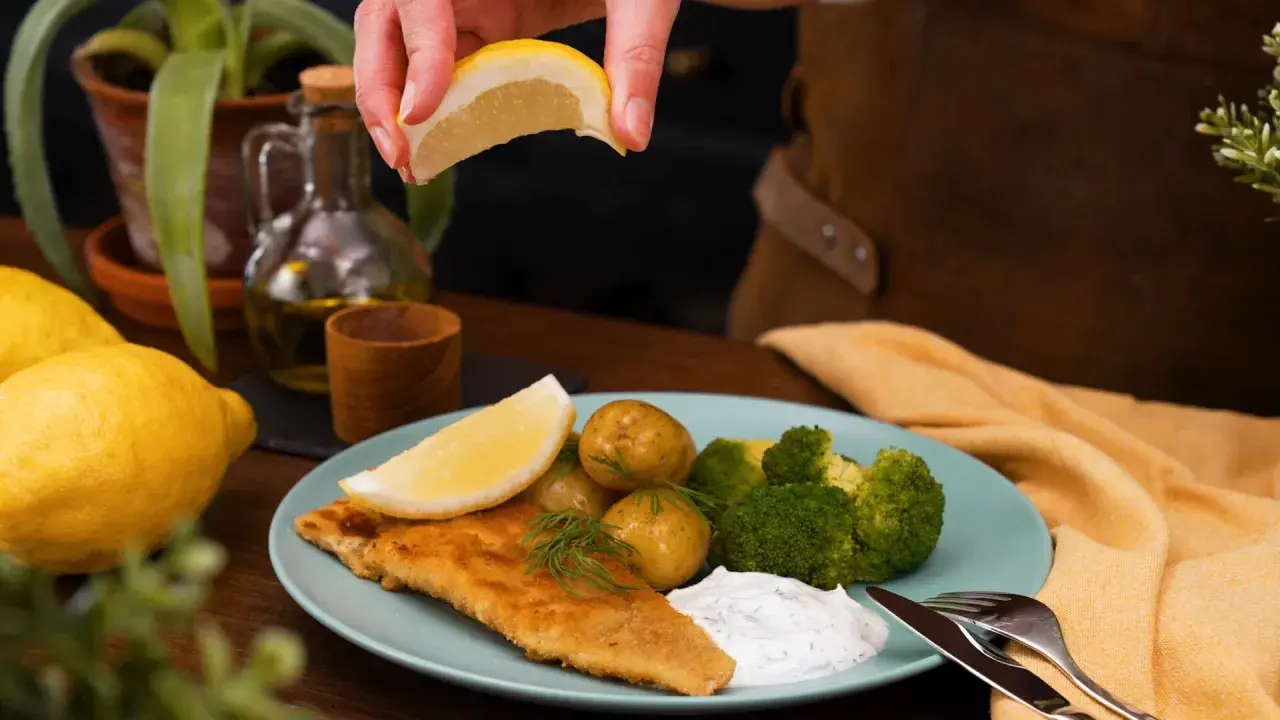 En saftig panerad fisk serveras med potatis, broccoli och en klick vit sås. En citronskiva pressas över fisken för extra smak.