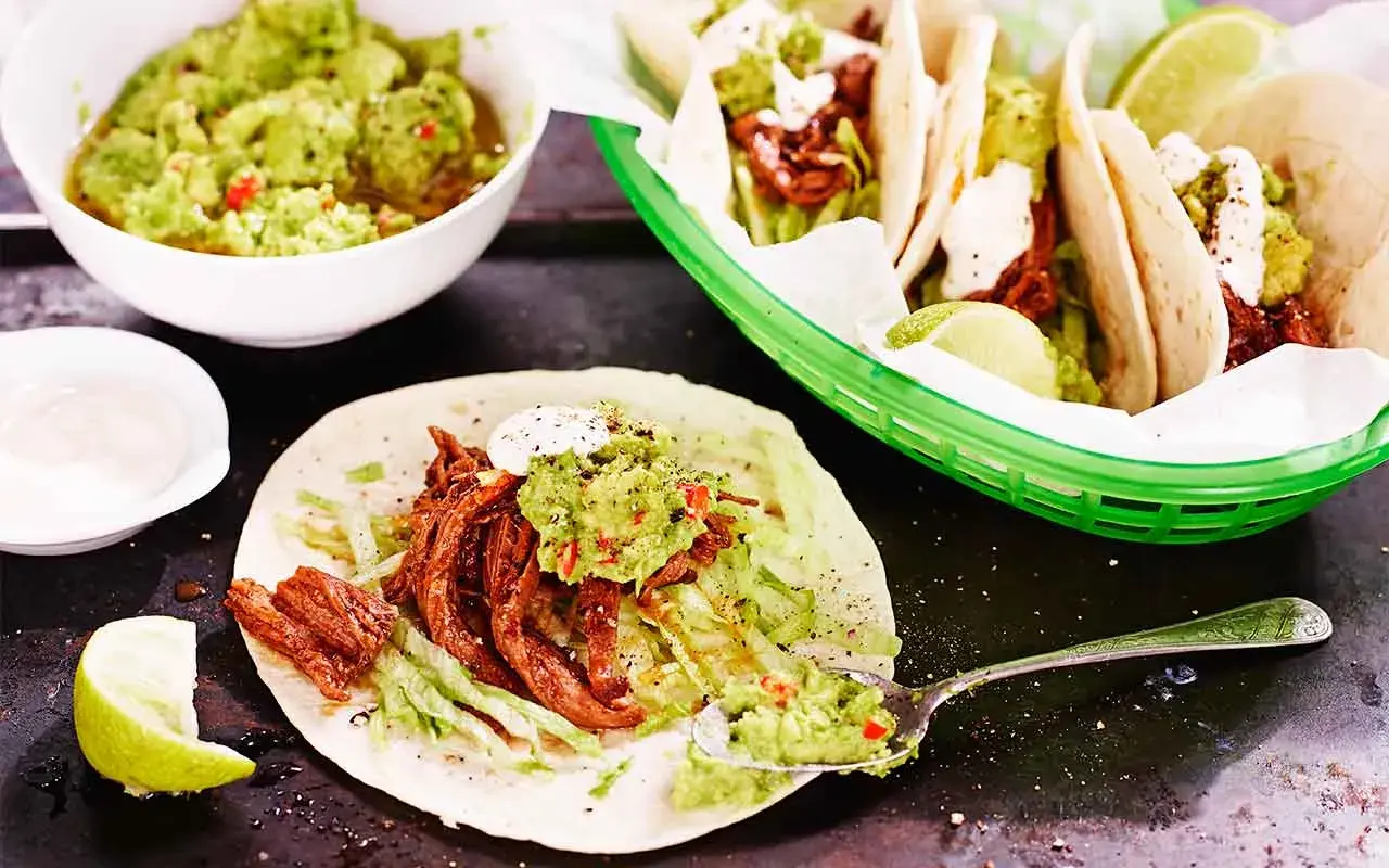 En läcker pulled beef taco med guacamole och strimlad sallad. En skål med mer guacamole och en limeklyfta syns också.