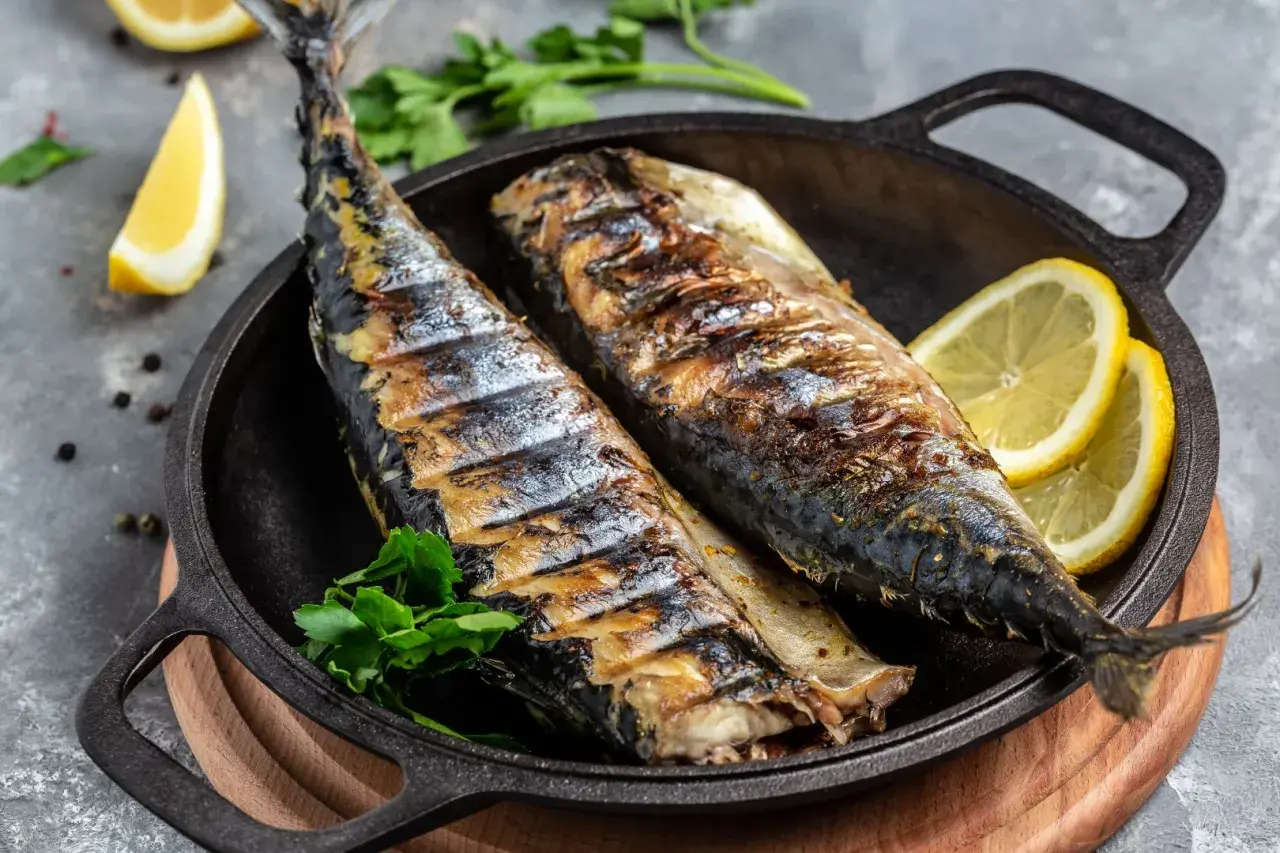 Två hela, grillade makrillar ligger i en gjutjärnspanna med citronklyftor och persilja.