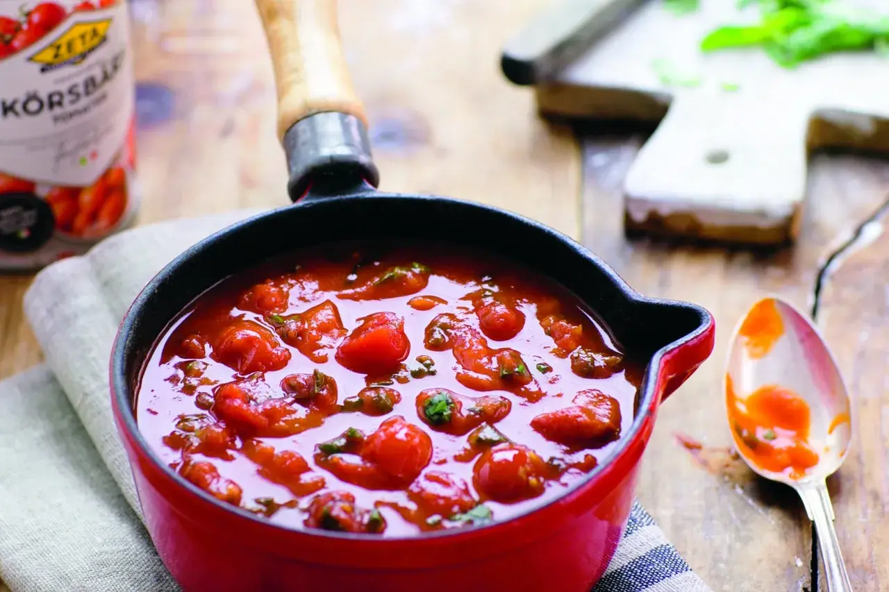 En röd kastrull fylld med en smakrik italiensk tomatsås med hela körsbärstomater och örter. En sked med sås ligger bredvid.