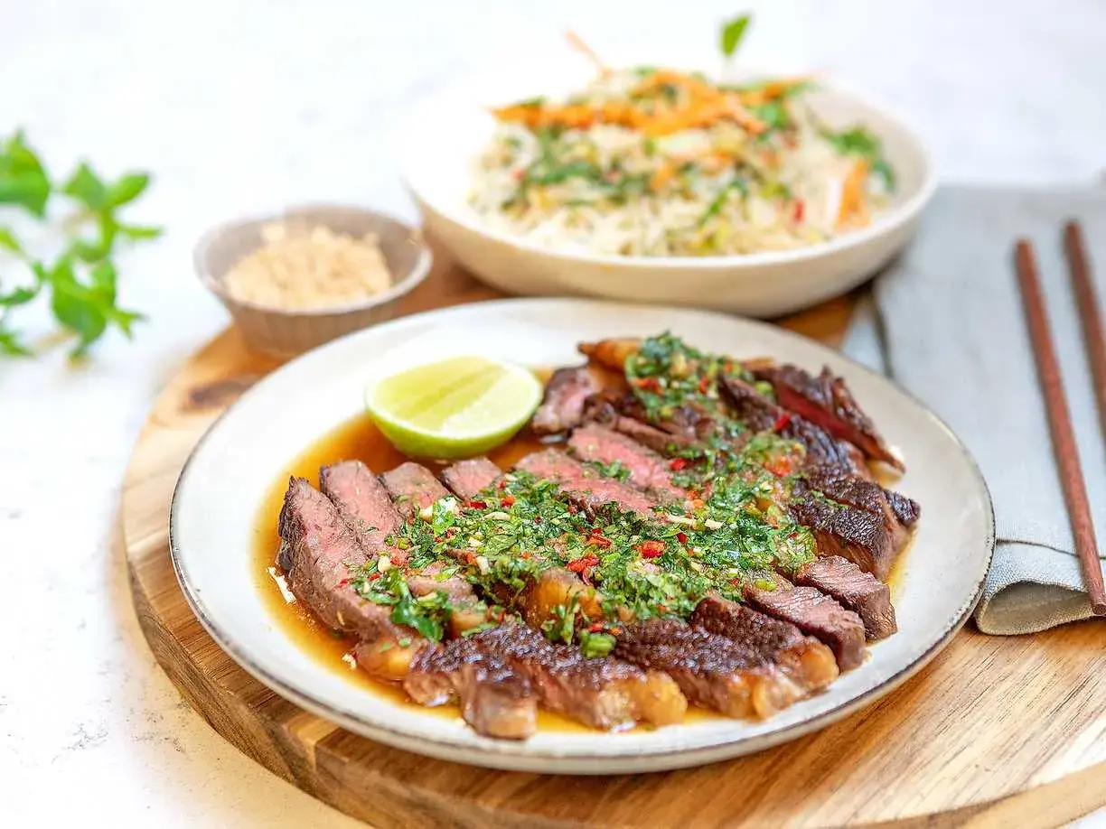 Skivad asiatisk entrecote med örtig sås, lime och en skål med ris.