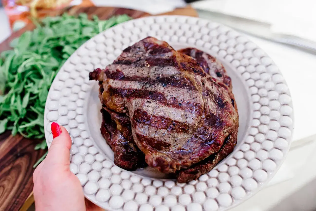 En hand med rött nagellack håller upp en tallrik med saftig biff tagliata, grillad till perfektion. Gröna blad syns i bakgrunden.