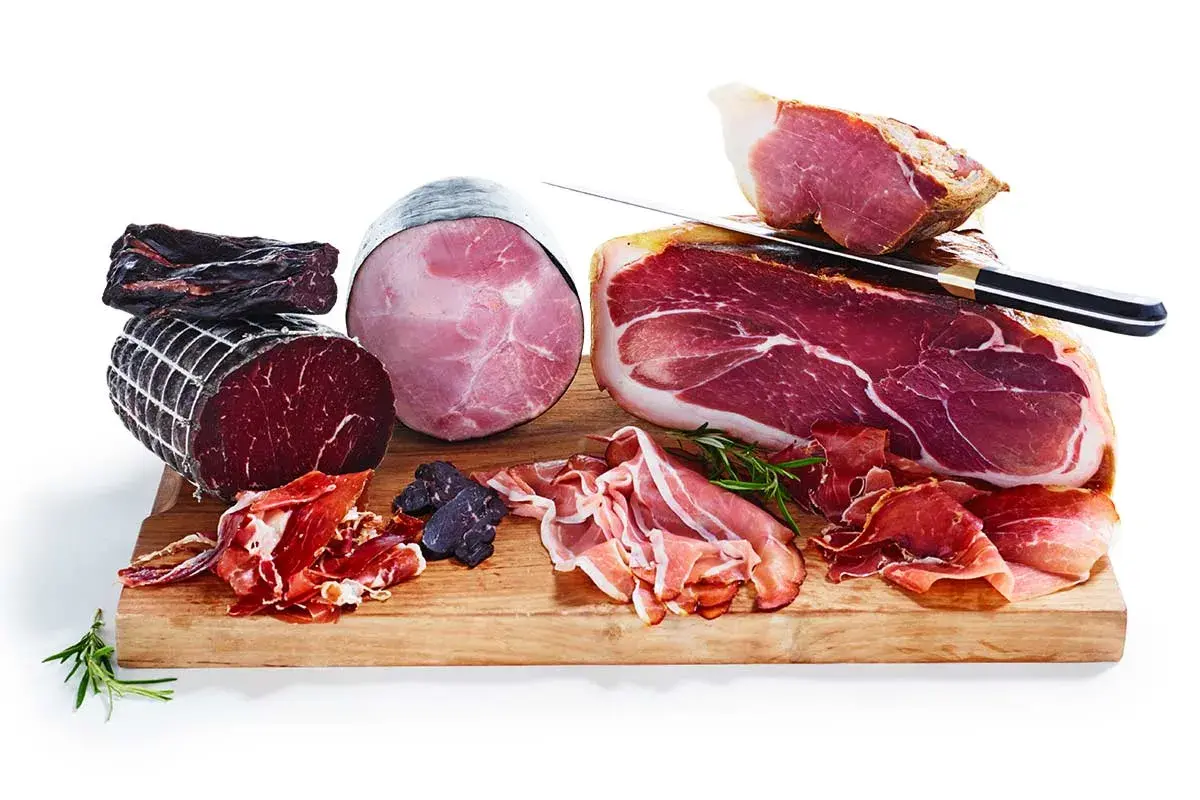 En träskärbräda med olika sorters charkuterier, inklusive en stor bit lufttorkad skinka, skivor av prosciutto och en rund, kokt skinka.