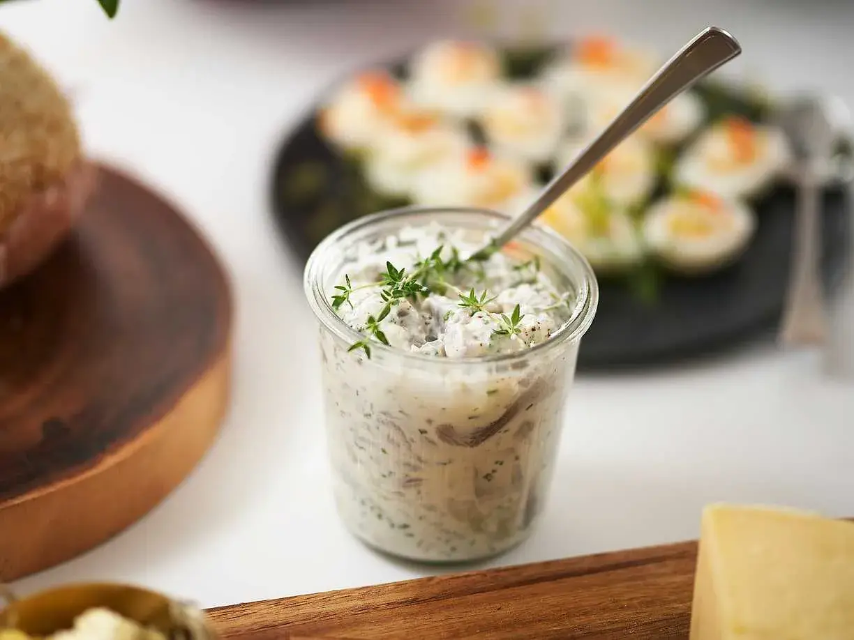 En burk med krämig wasabi sill, toppad med färska örter. I bakgrunden syns ägghalvor och en bit bröd.