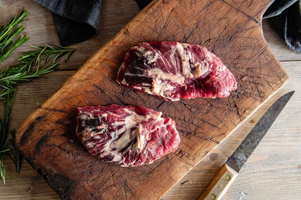 Två råa spider steak-biffar på en sliten träskärbräda, med rosmarin och en kniv.
