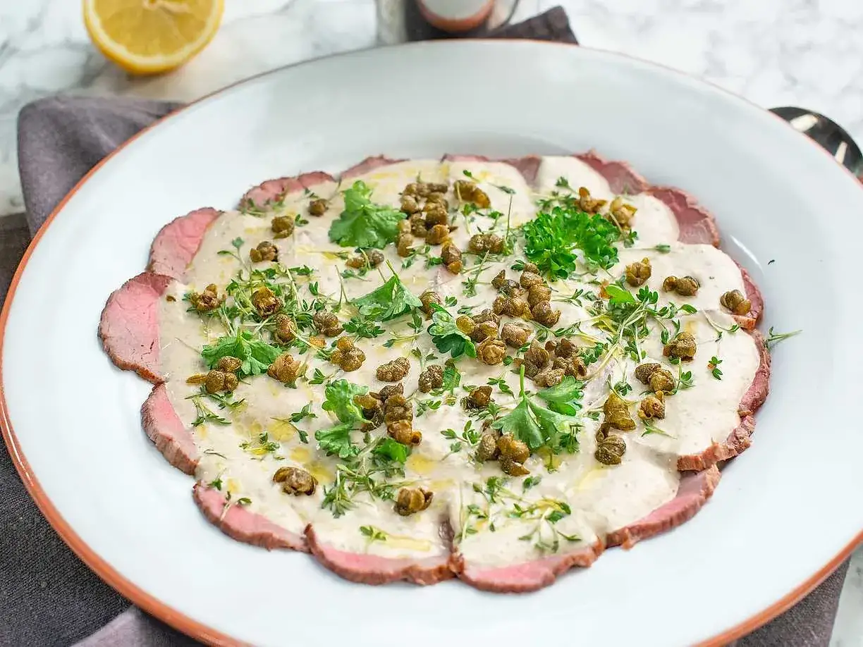 En tallrik med tunt skivad, rosa k&ouml;tt t&auml;ckt av en kr&auml;mig s&aring;s, kapris och f&auml;rska &ouml;rter. En klassisk vitello tonnato som ser uts&ouml;kt ut.