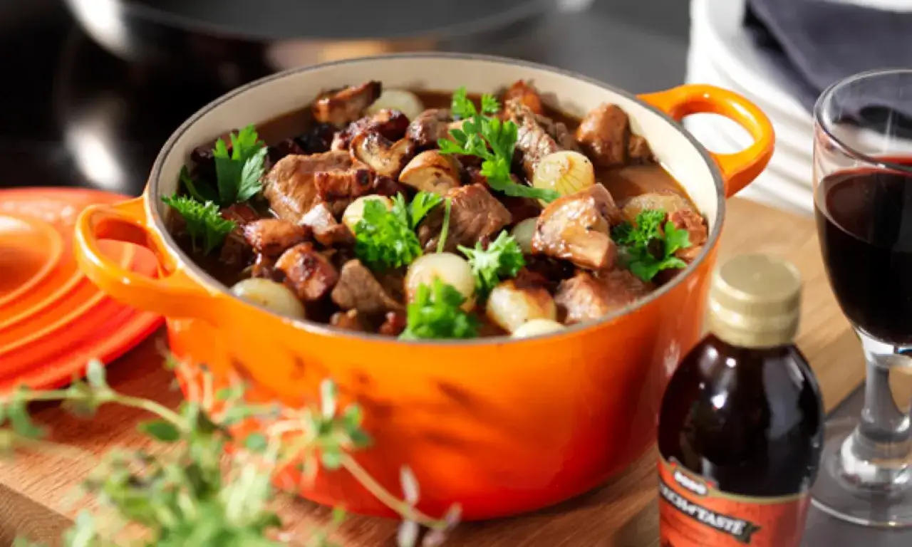 En mustig biff bourguignon i orange gryta, med champinjoner, p&auml;rll&ouml;k och persilja. Bredvid st&aring;r ett glas r&ouml;dvin och en flaska med smakf&ouml;rst&auml;rkare.