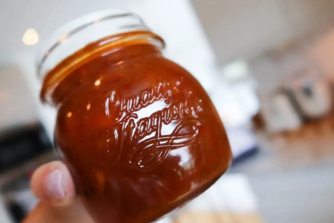 En hand håller ett glas med en mörkröd rabarber chutney. Texten