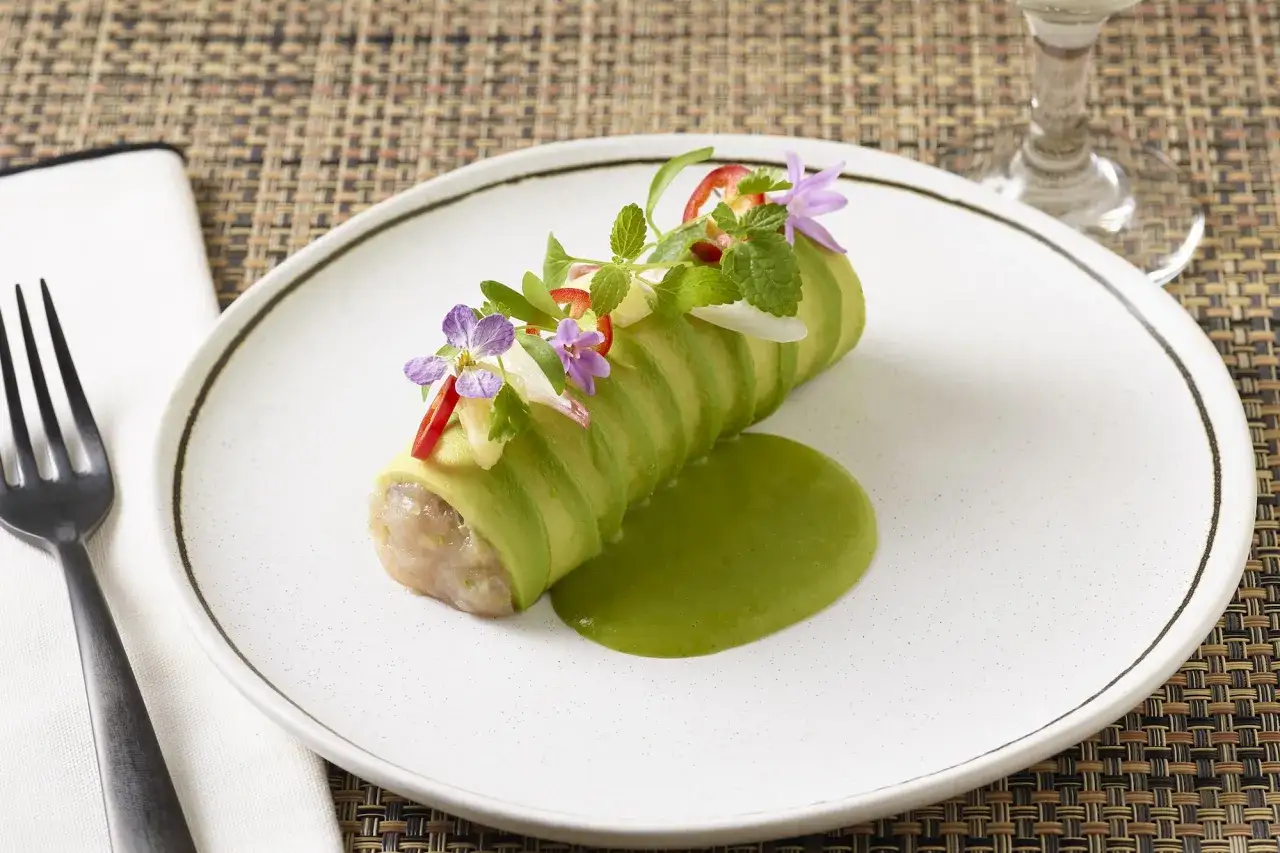 En konstn&auml;rlig presentation av tonfisk tartar insvept i skivor av kr&auml;mig avocado, toppad med &ouml;rter och blommor, serverad med en gr&ouml;n s&aring;s.