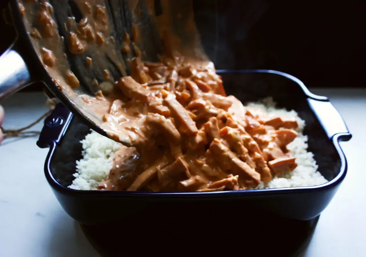 Slices of sausage in a creamy sauce are poured over white rice in a dark baking dish.