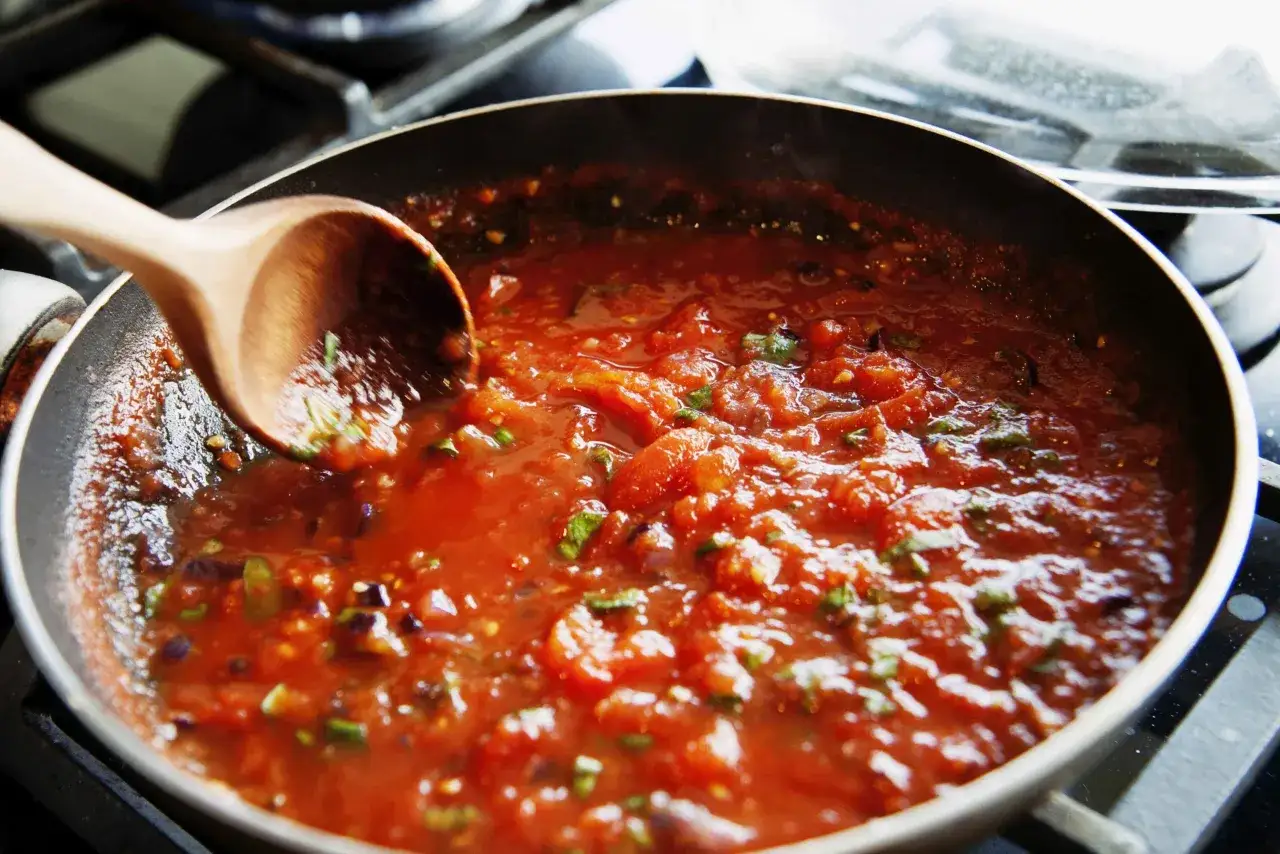 En tr&auml;sked r&ouml;r i en bubblande italiensk tomats&aring;s i en stekpanna. &Aring;nga stiger fr&aring;n den rika, r&ouml;da s&aring;sen med bitar av tomat och gr&ouml;na &ouml;rter.