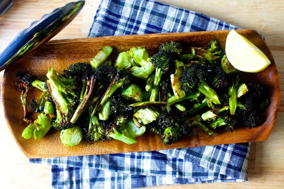 Rostad broccoli på ett träfat, perfekt som tillbehör eller som en del av ett enkelt broccoli recept. En citronklyfta ligger bredvid.
