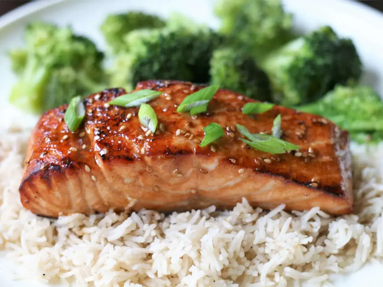 En saftig bit teriyaki lax serveras p&aring; en b&auml;dd av ris, toppad med salladsl&ouml;k och sesamfr&ouml;n, med &aring;ngad broccoli i bakgrunden.