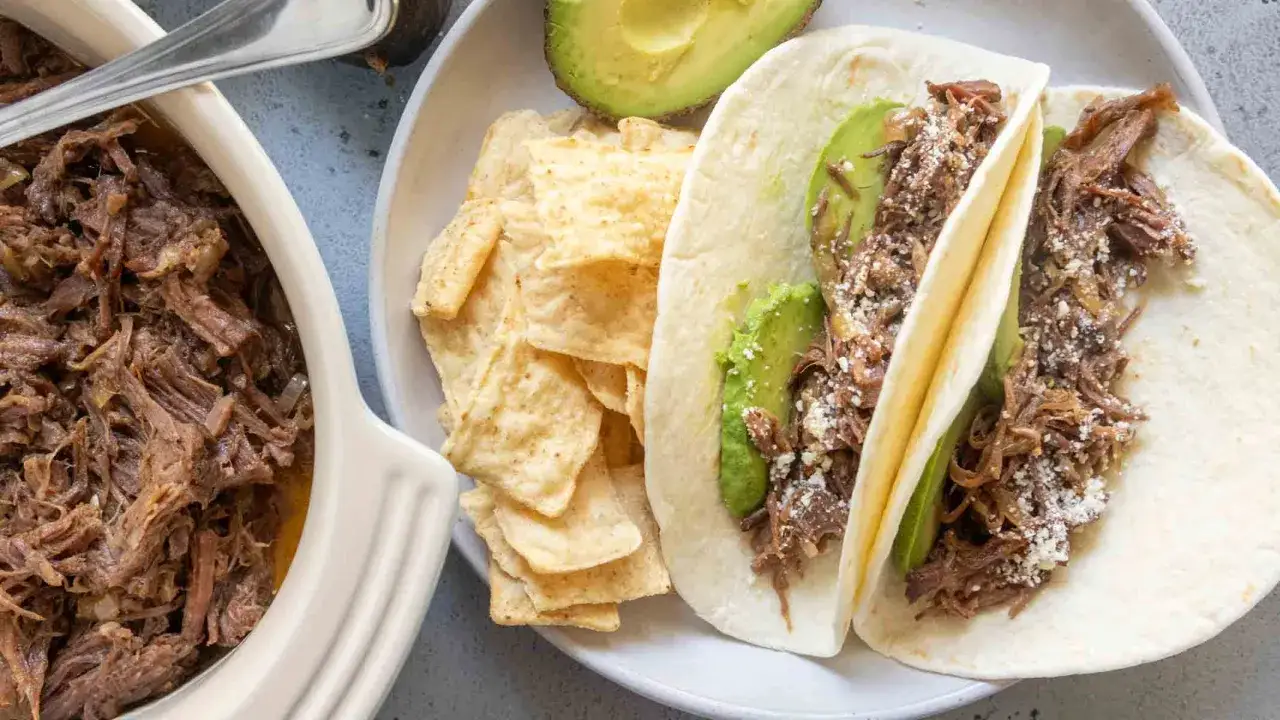 Två pulled beef tacos med avokado och ost, serverade med tortillachips och en skål med mer saftigt pulled beef.