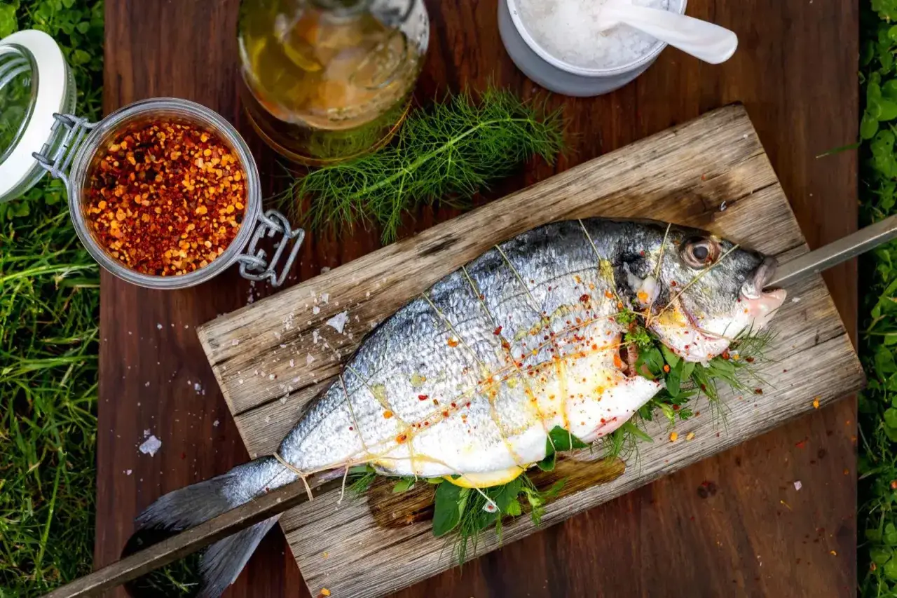 En hel, grillad fisk på ett träspett, redo att tillagas. Kryddad med chili och örter, med citron och olja bredvid.