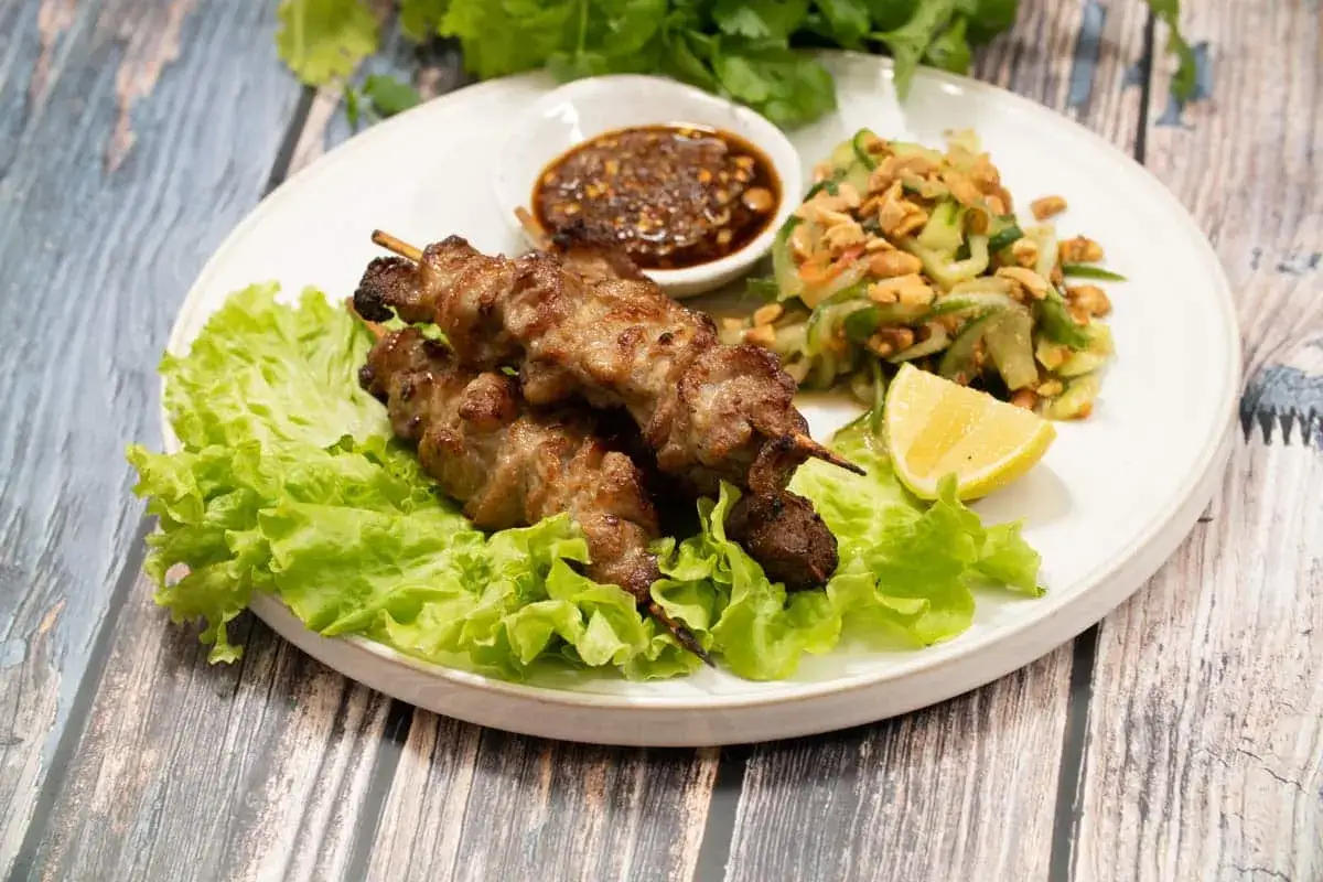 Grillade spett med fläsk, kända som moo ping, serveras med en fräsch gurksallad, dippsås och en citronskiva.