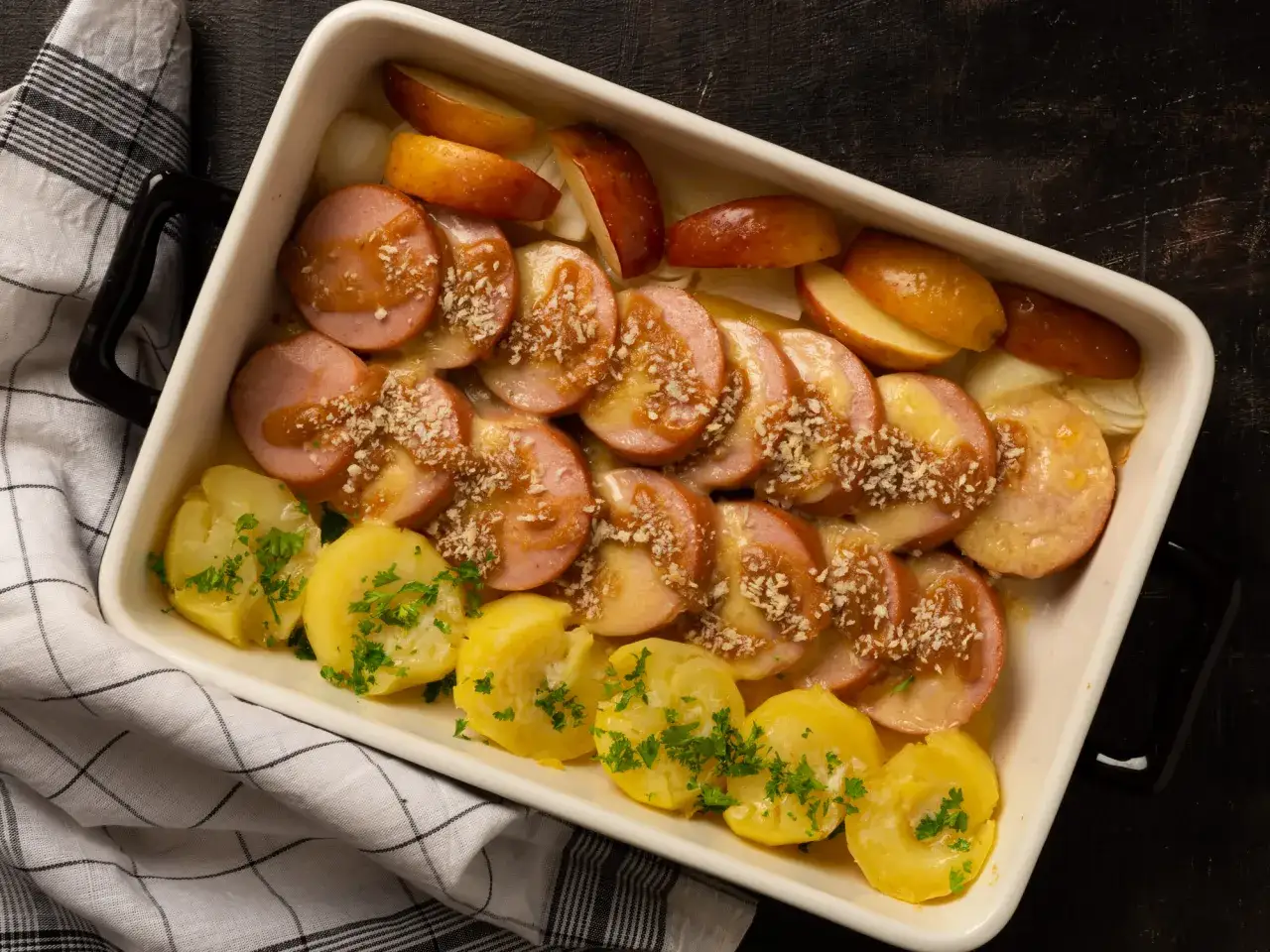 Ugnsgratinerad falukorv med potatis och äpple i en vit ugnsform. Ströbröd och persilja toppar rätten.