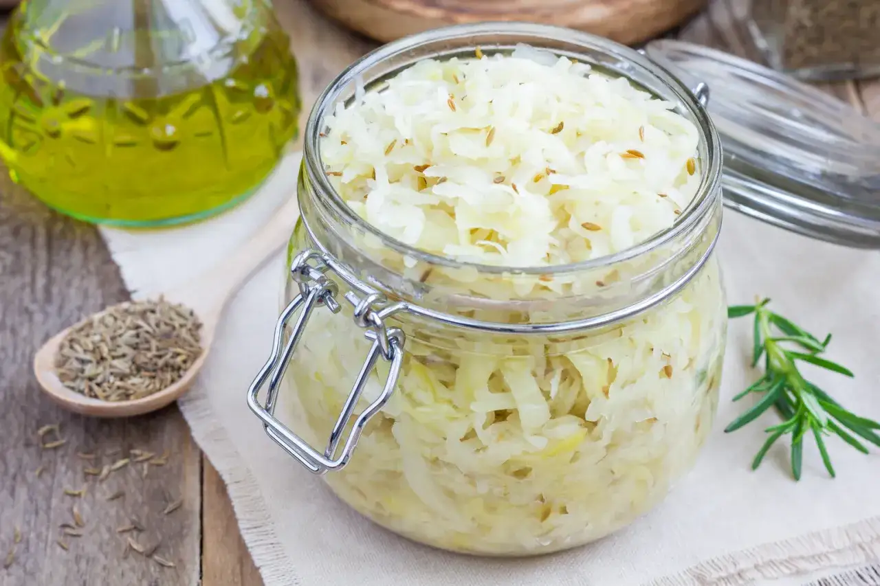 En burk med hemlagad surkål, perfekt för ditt surkål recept. En sked med kumminfrön och en flaska olja syns bredvid.
