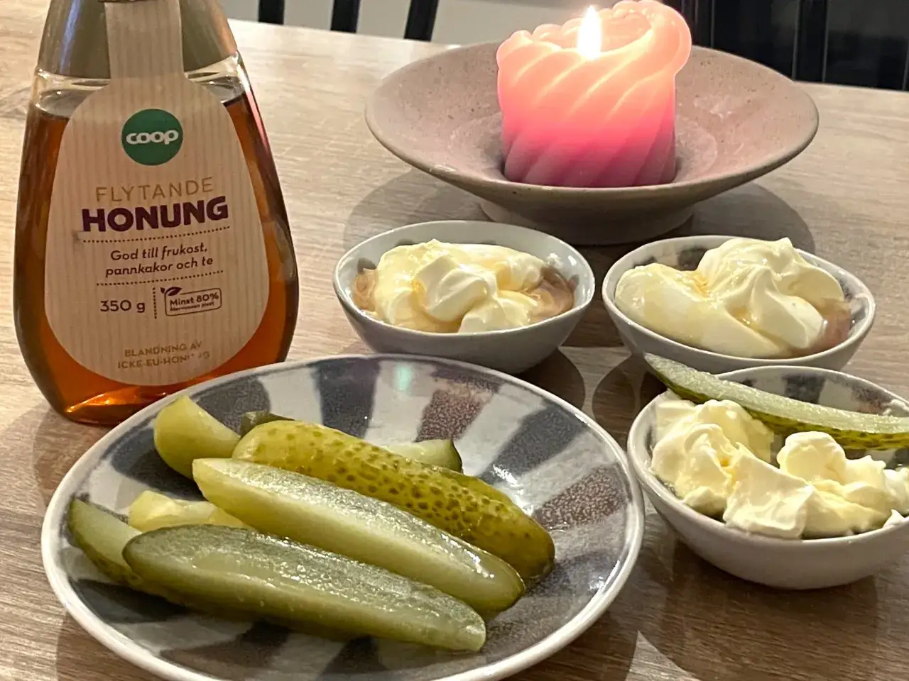 Saltgurka, smetana och honung p&aring; bordet, med en t&auml;nd ljuslykta i bakgrunden. En flaska honung st&aring;r till v&auml;nster.