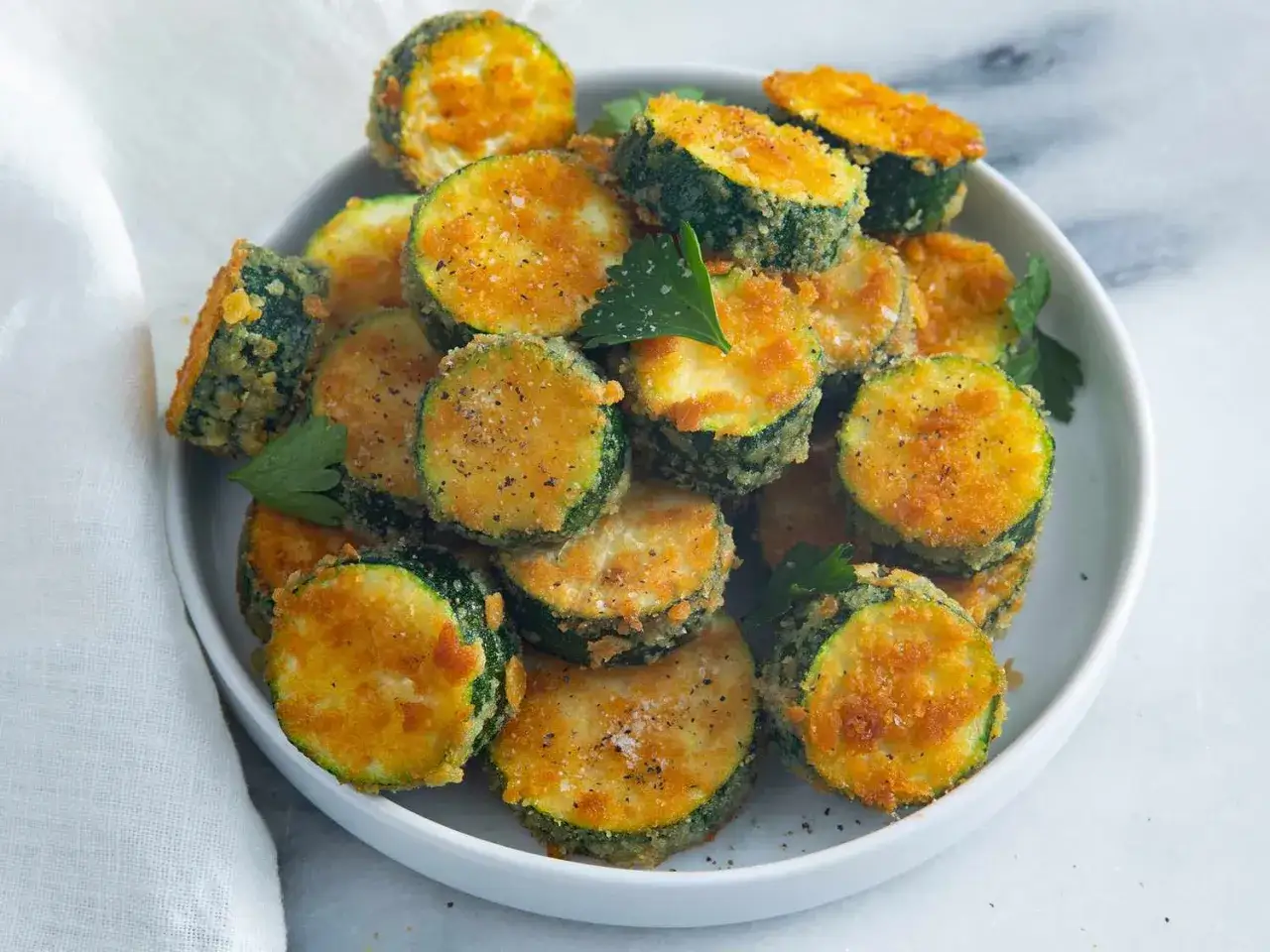 Krispiga, gyllene skivor av stekt zucchini, toppade med salt och peppar, serveras på en vit tallrik med persilja.
