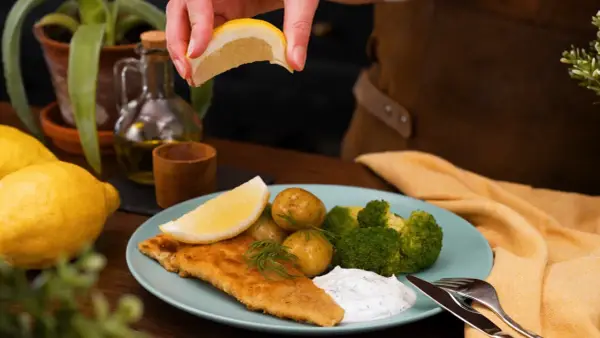 En saftig panerad fisk serveras med potatis, broccoli och en klick vit sås. En citronskiva pressas över fisken för extra smak.